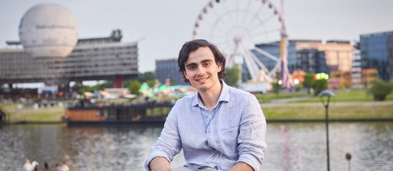Man seated outside with a river, ferris wheel and buildings in background.