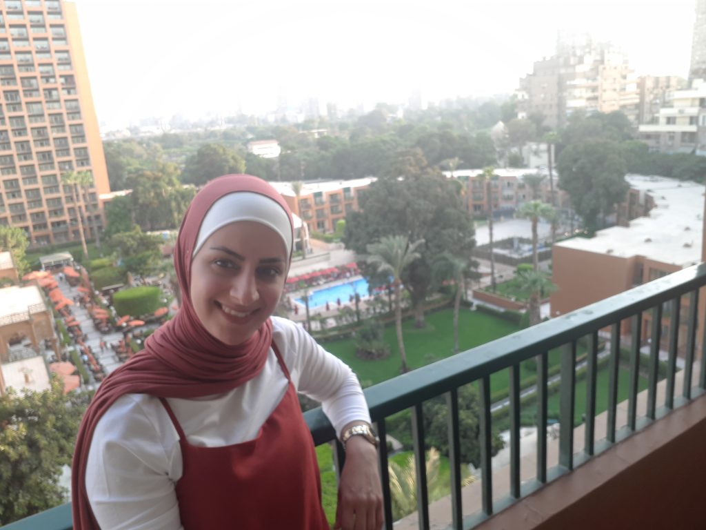 A smiling woman in red headscarf stands on a balcony with a nice garden view