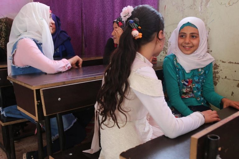 Foreground of photographs shows two young girls seated at a desk facing each other. The girl to the right is wearing a white headscarf and green top. To the left the girl has long dark hair in a ponytail and is wearing a white top.