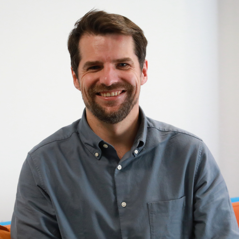 A man with short brown hair and a beard, smiling, wears a buttoned-up gray shirt. He is sitting indoors against a plain white background.