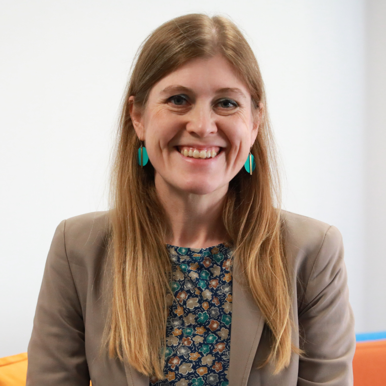 A woman with long blonde hair smiles at the camera. She is wearing a tan blazer, a patterned blouse, and turquoise earrings. The background is plain and light-colored.