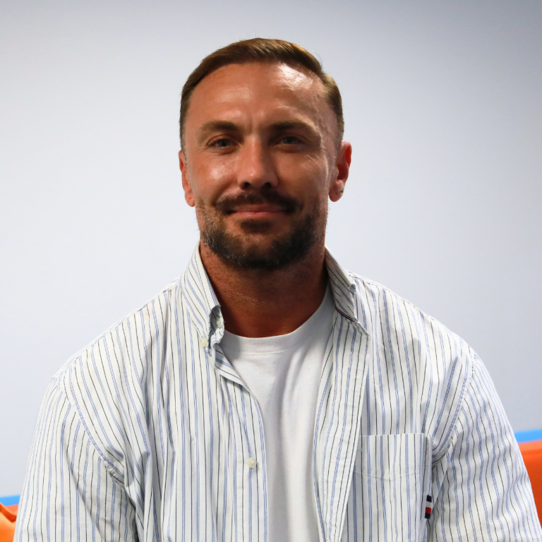 A man with short brown hair and a beard wears a white t-shirt and a white striped button-up shirt, sitting indoors against a light blue wall.