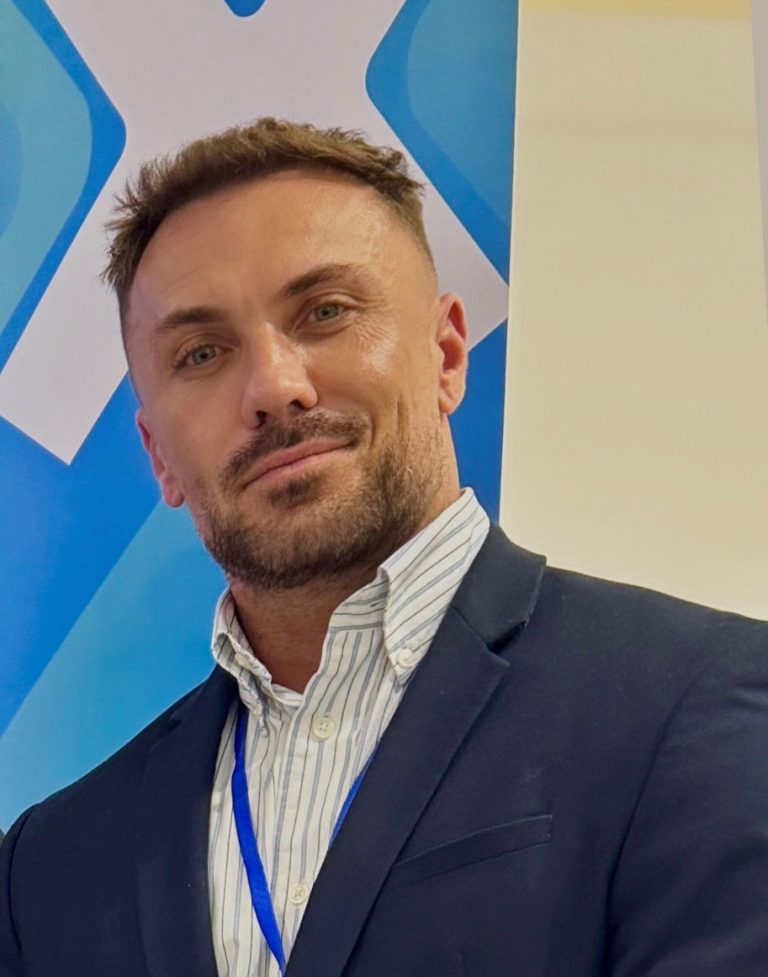 A man with short brown hair and a trimmed beard, wearing a dark suit jacket, striped shirt, and blue lanyard, stands in front of a blue and white background.