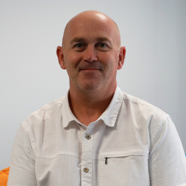 A middle-aged man with a shaved head, wearing a white button-up shirt, smiles while sitting in front of a plain light-colored background.