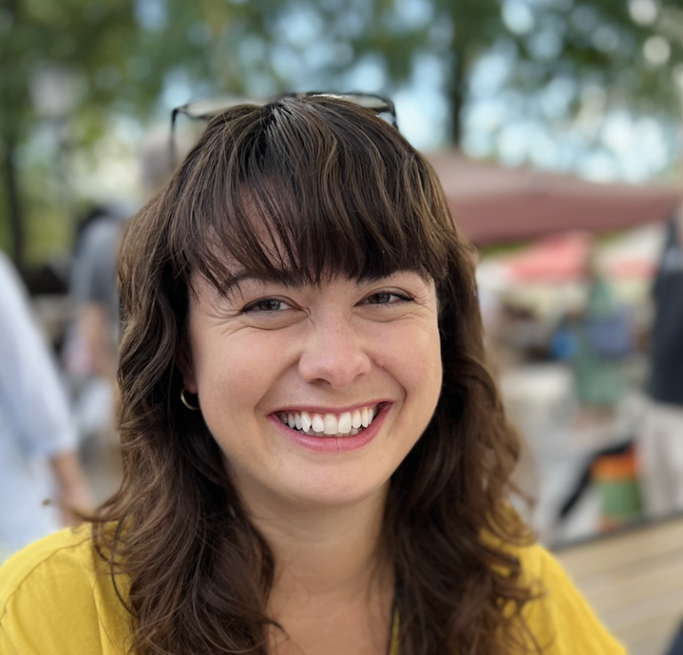 A woman with wavy brown hair and bangs smiles brightly while sitting outdoors. She is wearing a yellow top and there are blurred people and greenery in the background.
