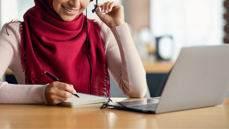 A woman wearing a red hijab and headset smiles while taking notes in a notebook and looking at her laptop on a desk.