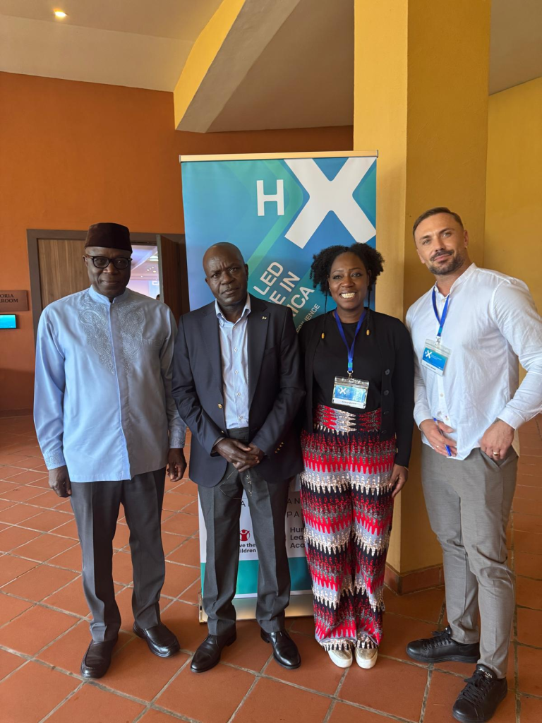 Four people stand indoors in front of a blue HX Lead Africa banner, smiling for the camera. The group includes three men in business attire and a woman wearing a colorful outfit and conference badge.