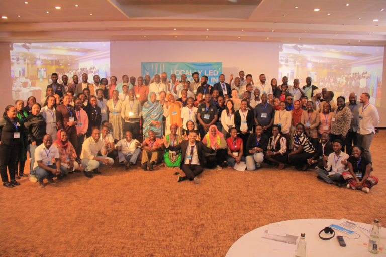 A large group of people pose together for a group photo in a conference hall, with a banner in the background reading Locally Led. The group is diverse and smiling, and the room is brightly lit.