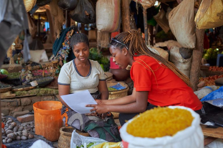 Two women sit in a busy market surrounded by produce and goods. One, in a red shirt, shows the other a piece of paper—possibly a Humanitarian AI report—as they discuss attentively amid baskets and bags of food.