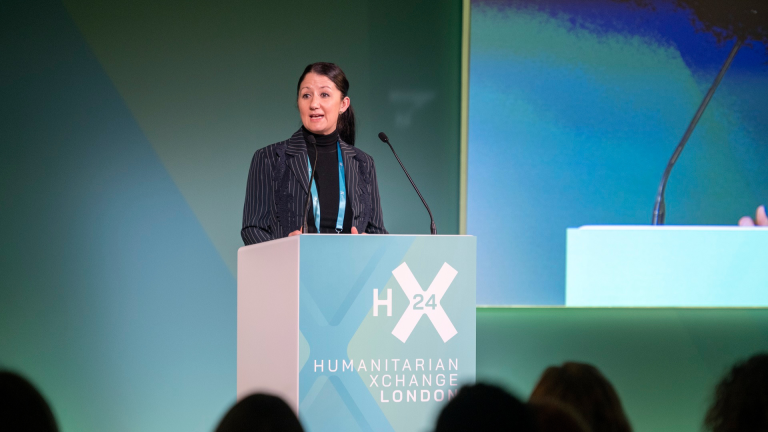 A woman speaks at a podium labeled H24 Humanitarian Xchange London on a stage with a green backdrop. She is wearing a dark pinstripe blazer and black turtleneck, addressing an audience.