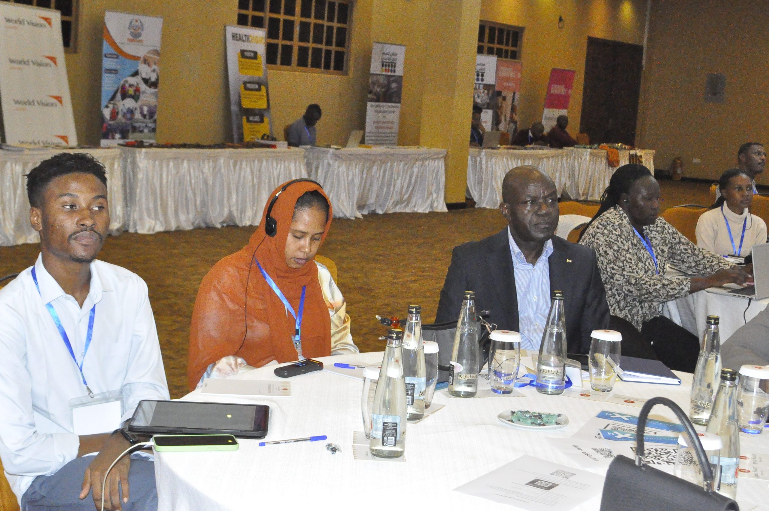 A group of people sit around a round table at a conference, with name tags and bottled water on the table. Some attendees are listening attentively; banners and more participants are visible in the background.