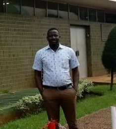 A man wearing a light blue patterned shirt and brown pants stands on a path in front of a green-tiled building, surrounded by grass and plants.