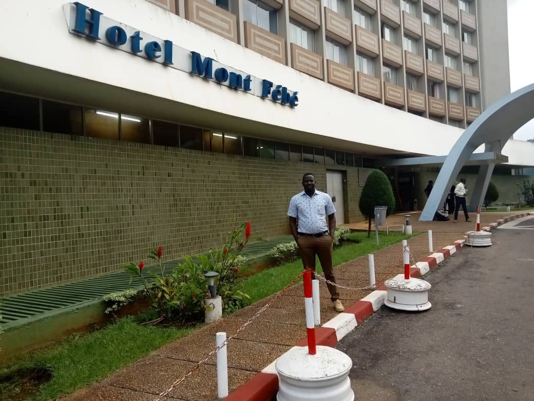 A man stands smiling in front of the Hotel Mont Fébé building, near a flower bed and walkway, as Learning with Kaya meet Julius from Nigeria; the hotel sign is visible above and part of the entrance appears on the right.