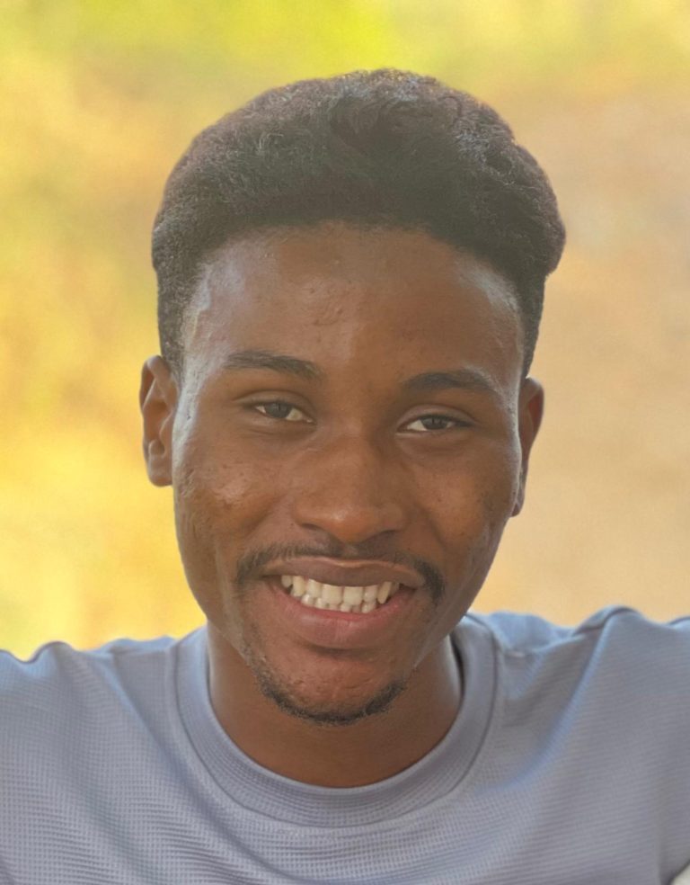 A young man with short black hair and a trimmed mustache and beard smiles at the camera. He is wearing a light gray shirt and is outdoors with a blurred, sunlit background.