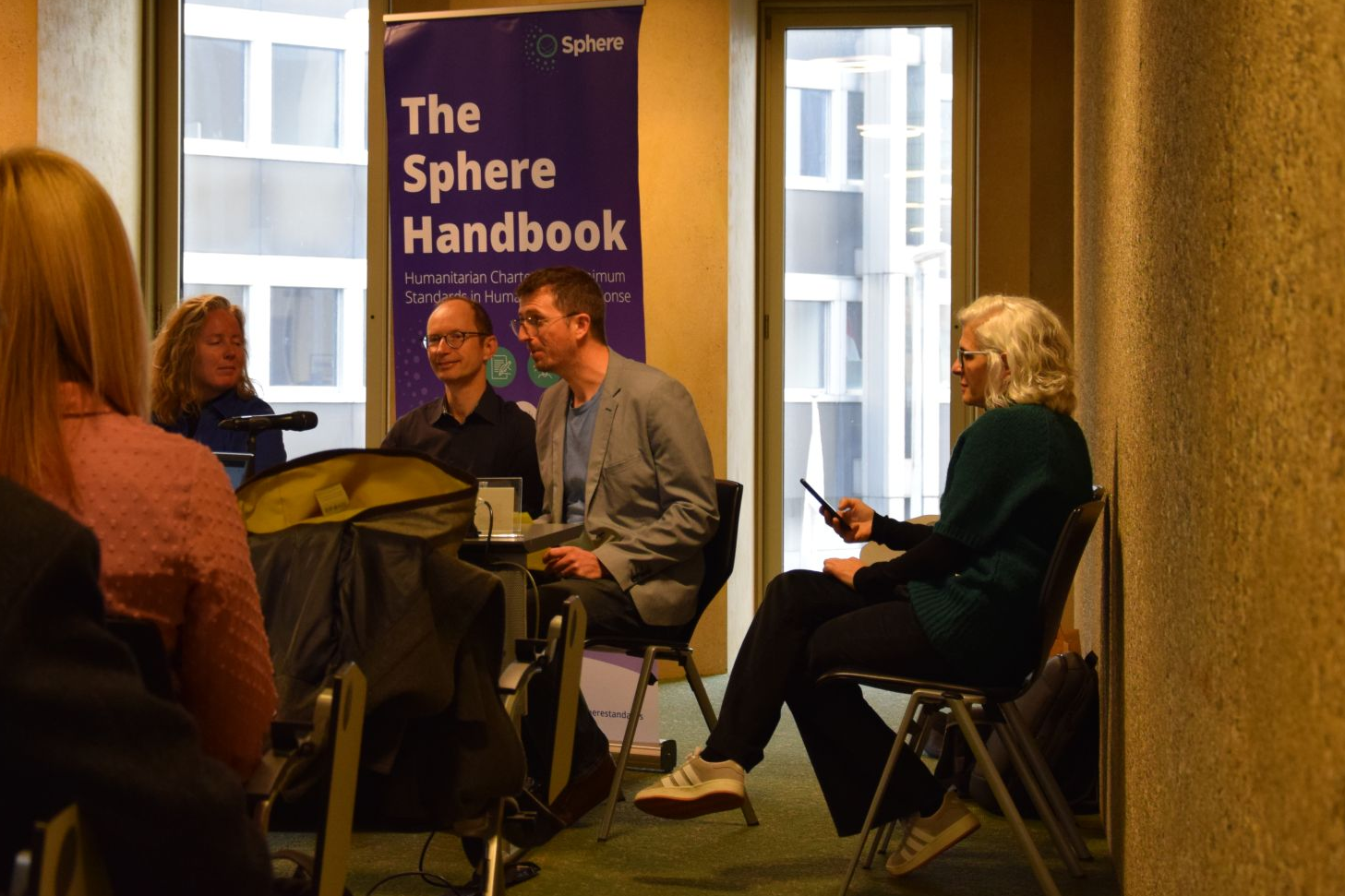 People sit in a small conference room listening to a panel of four speakers in front of a banner reading The Sphere Handbook. One panelist is speaking while others listen, and attendees are seated facing them.