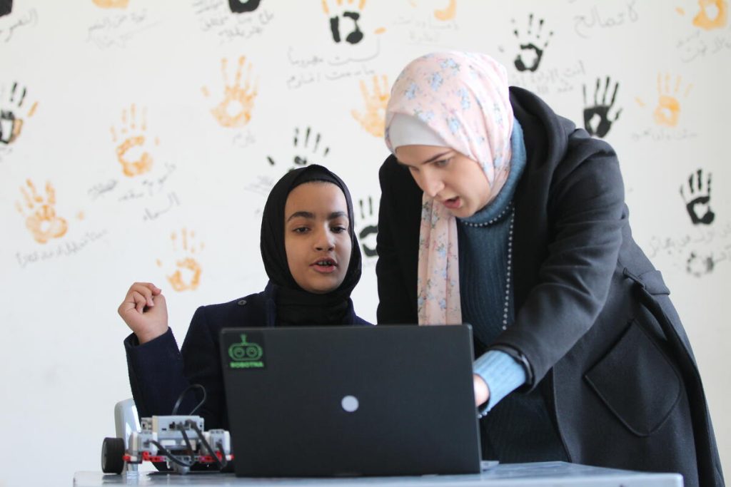 Two women wearing hijabs work together at a laptop. One is seated, the other leans over, pointing at the screen. A small robot is on the table. Behind them is a wall with colorful handprints and Arabic writing.