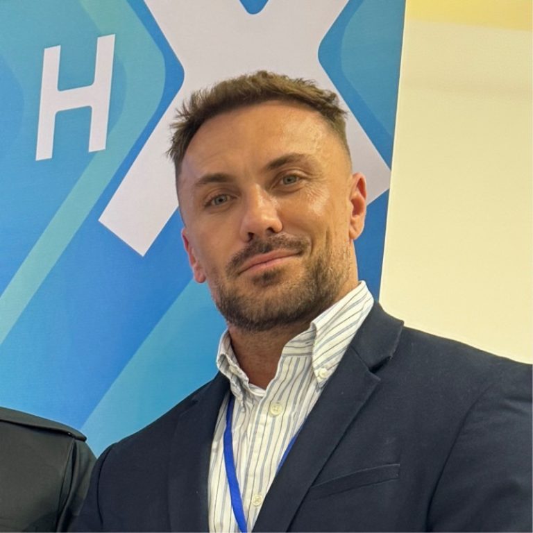 A man with short brown hair and trimmed beard, wearing a navy blazer and striped dress shirt, stands in front of a blue background with white letters and shapes, smiling slightly at the camera.