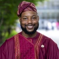 A man smiling outdoors, wearing a traditional maroon and gold African attire with a matching cap. There are green trees and blurred people in the background.