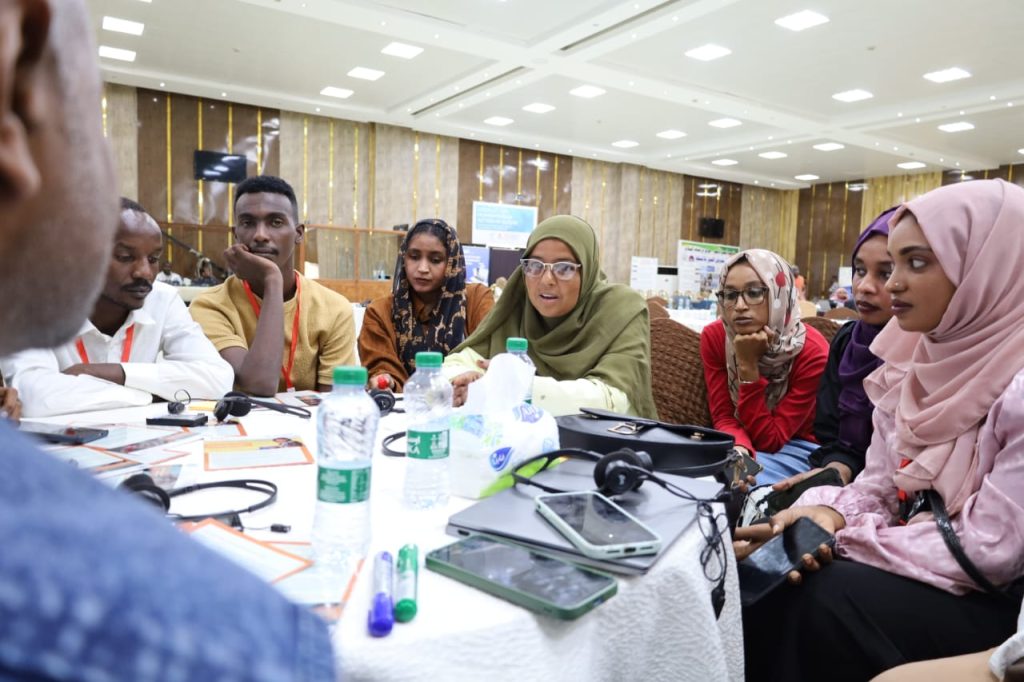 A group of people sit around a round table in a conference room, engaged in discussion about HX Port Sudan - locally led action in the Sudan response. Water bottles, notepads, and electronic devices are on the table as attendees focus attentively.
