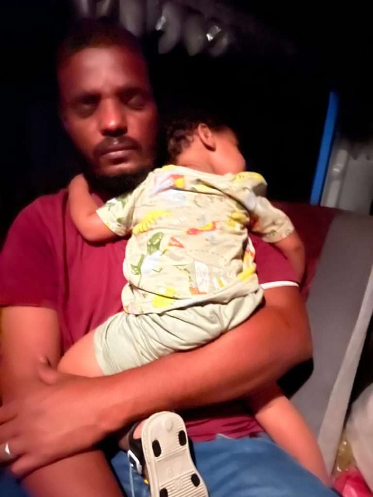A man sits with his eyes closed, holding a sleeping child in his arms. The child is resting comfortably against his chest, wearing a patterned shirt and shorts. The background appears dimly lit.