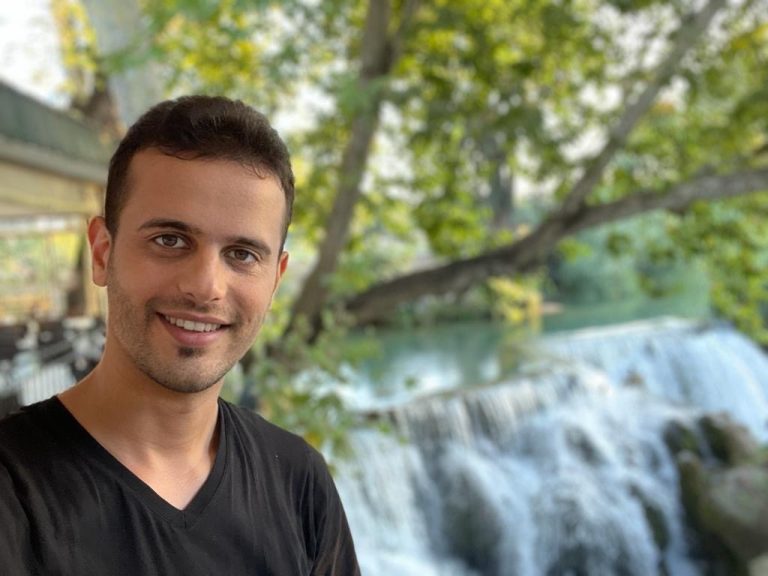 A man with short dark hair and a black shirt smiles at the camera while standing outdoors near a flowing waterfall and lush green trees.