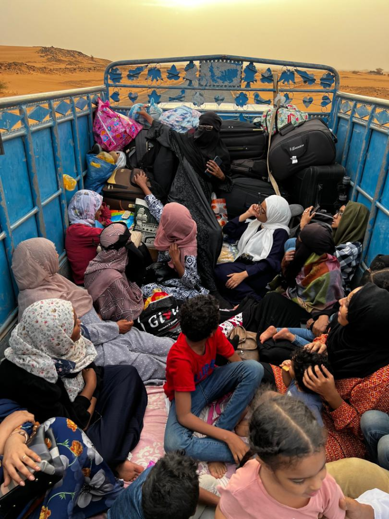 A group of women and children, some wearing headscarves, sit closely together in the back of a blue open truck filled with suitcases, bags, and belongings, set against a sandy desert landscape at sunset.