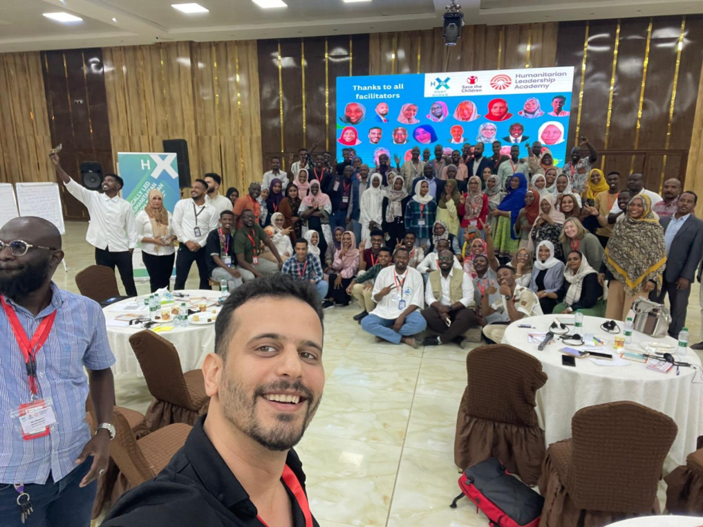 A man takes a selfie in front of a large, diverse group of people posing and smiling in a conference room. Behind them is a screen showing faces and logos, and tables with event materials are visible.