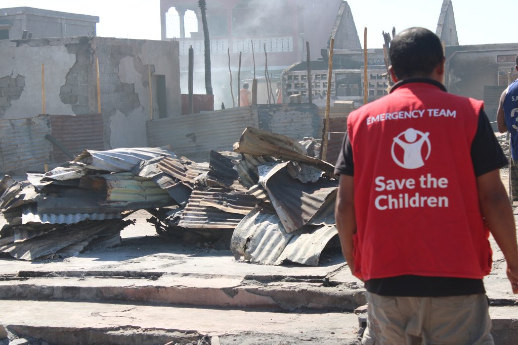 A person wearing a red Save the Children Emergency Team vest stands in front of destroyed buildings and debris, with smoke rising in the background, suggesting recent devastation or disaster.