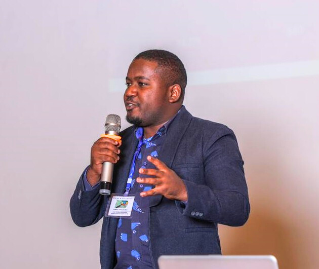 A man wearing a navy blazer and blue patterned shirt speaks into a microphone while gesturing with one hand, likely hosting the Fresh Humanitarian Perspectives podcast on humanitarian AI. He has a conference badge around his neck and stands in front of a light background.