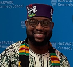 A smiling man in traditional African attire and hat, wearing glasses and a colorful kente stole, stands in front of a blue Harvard Kennedy School backdrop.