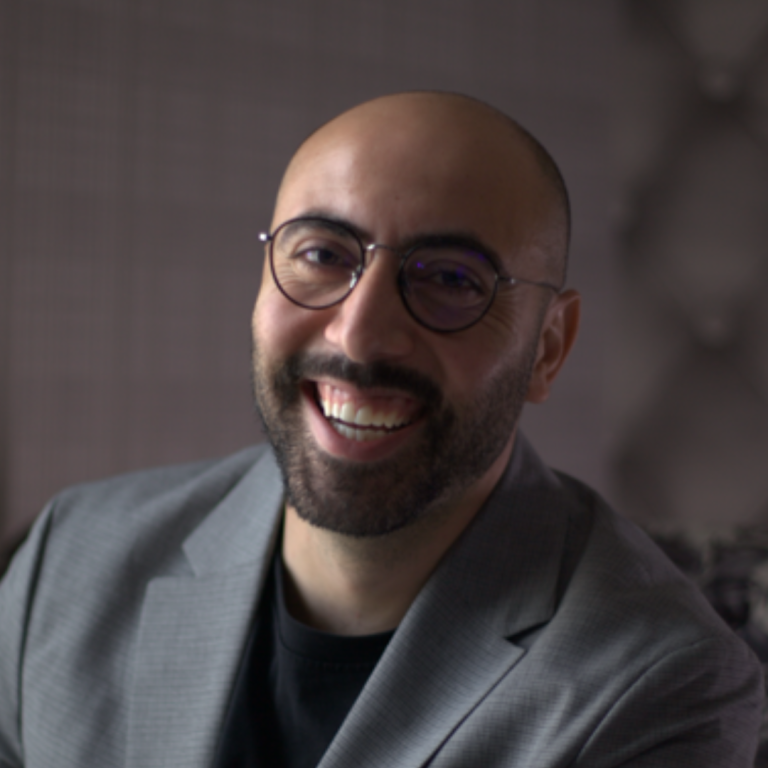 A smiling man with a beard and glasses, wearing a gray blazer over a black shirt, is sitting indoors with a softly blurred background.
