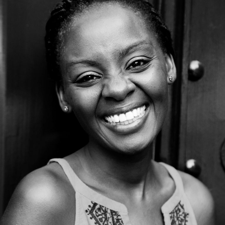A woman with braided hair smiles broadly, showing her teeth. She wears stud earrings and a sleeveless top with a decorative pattern. The photo is in black and white, with a blurred dark background.