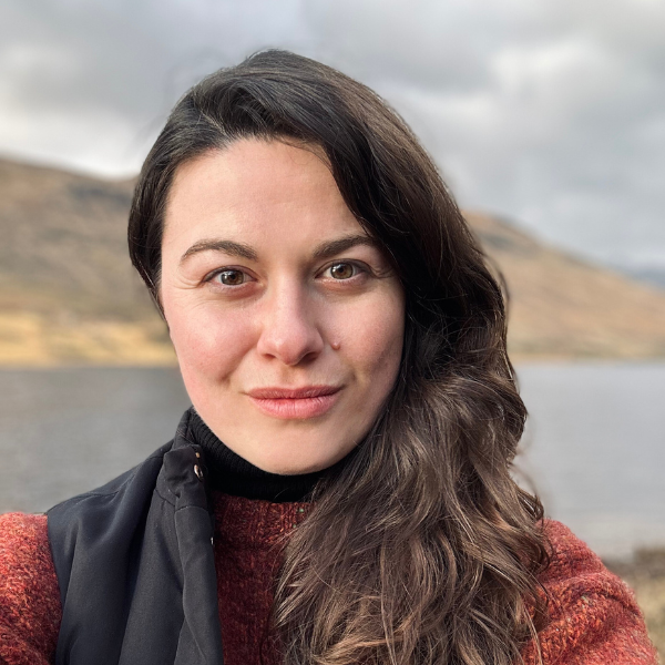 A woman with long brown hair and a slight smile takes a selfie outdoors near a lake, with hills and cloudy skies in the background. She wears a rust-colored sweater and black vest.