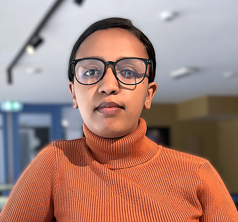 A woman wearing glasses and an orange ribbed turtleneck sits at a table with her hands clasped, looking at the camera. The background shows a modern, brightly lit room with blue and beige walls.