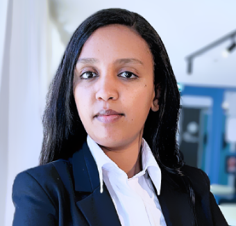 A confident woman with long black hair, wearing a black blazer and white shirt, stands with arms crossed in a modern, brightly lit office space.