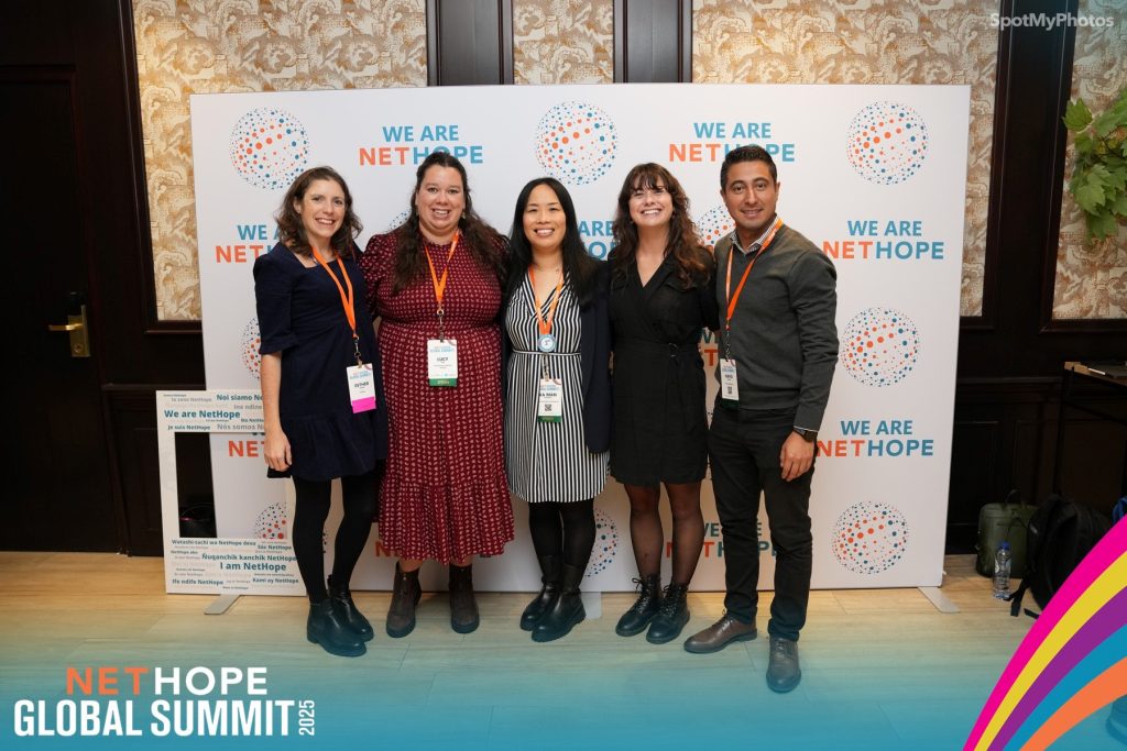Five people stand side by side, smiling at the camera, in front of a We Are NetHope backdrop at the NET HOPE Global Summit 2023. All wear conference badges and orange lanyards.