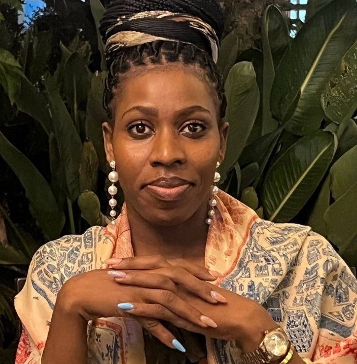 A woman with braided hair styled in an updo, wearing pearl earrings and a patterned shawl, sits with her hands folded on a table. She has blue nail polish and a gold watch. Lush green plants are in the background.