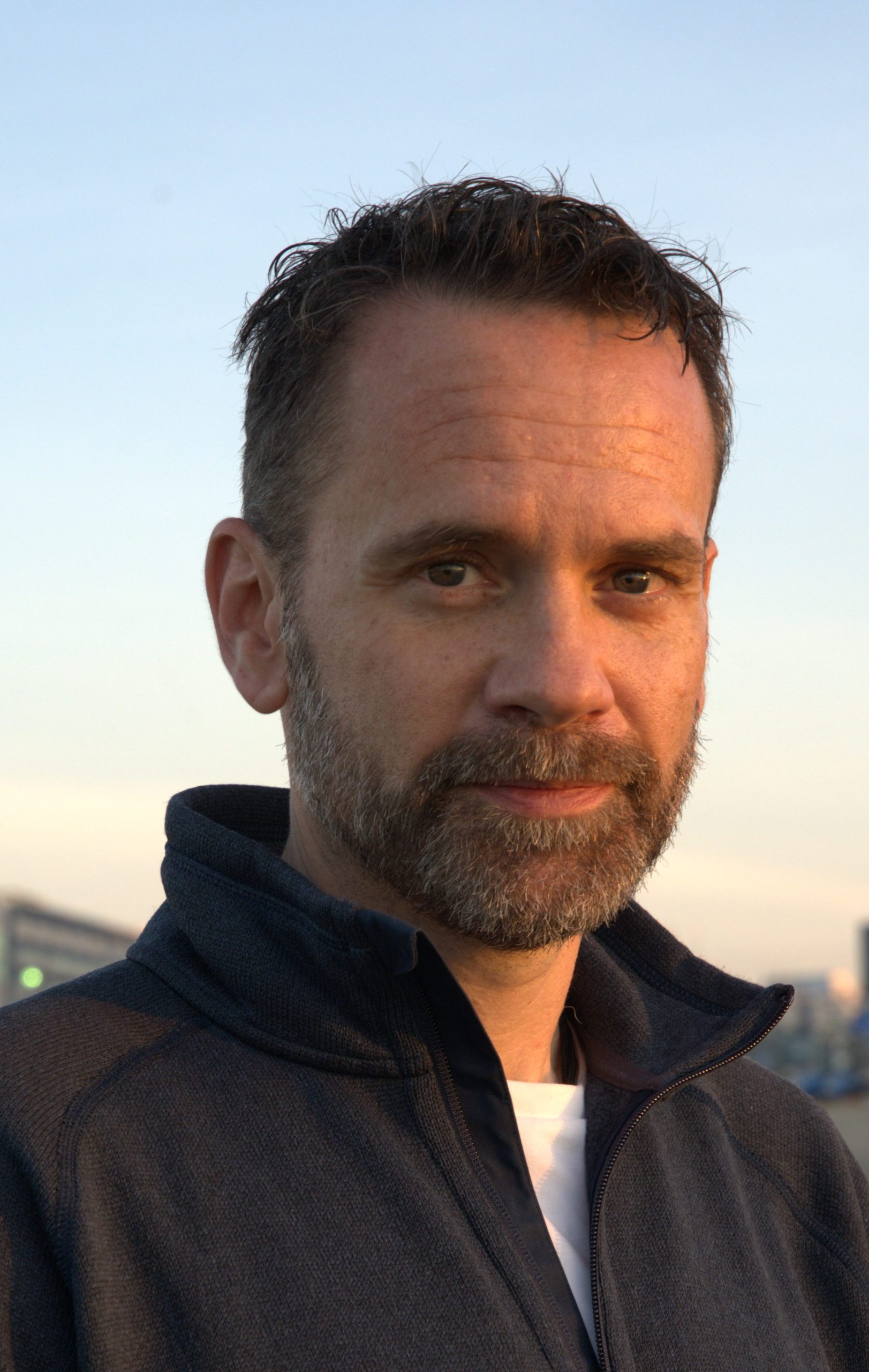 A man with short dark hair and a beard, wearing a dark jacket over a white shirt, stands outdoors at sunset with buildings and a blue sky in the background.