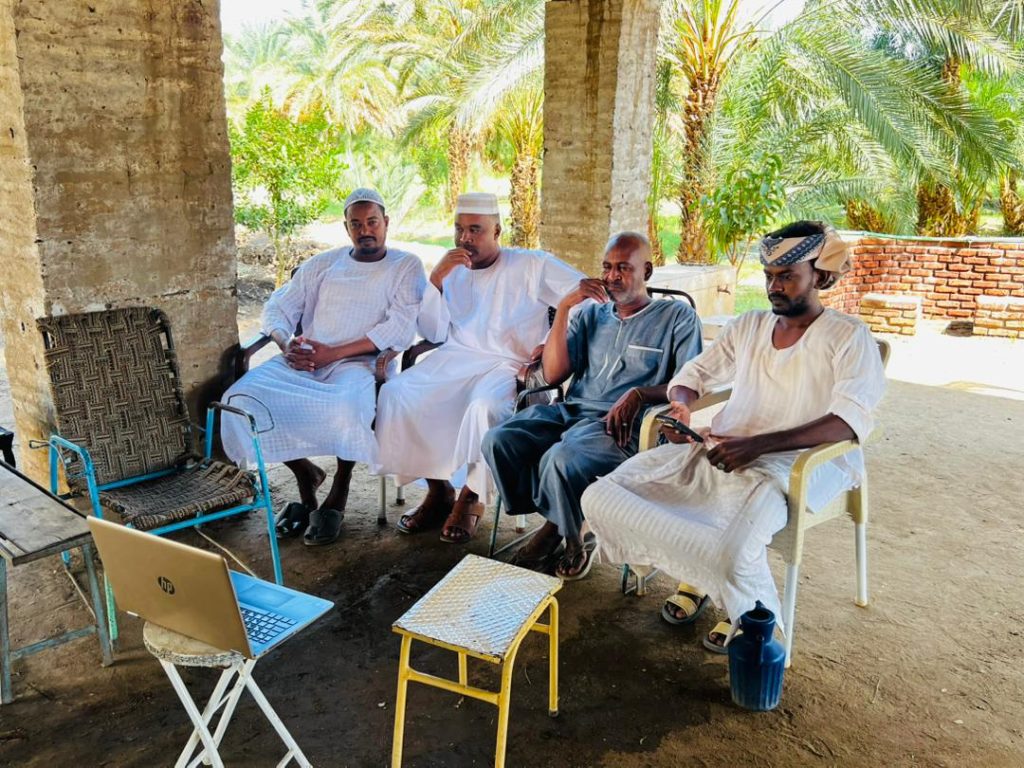 Four men in traditional clothing sit on chairs outdoors, under a shaded structure, watching a laptop on a small table. Palm trees and sunlight are visible in the background.