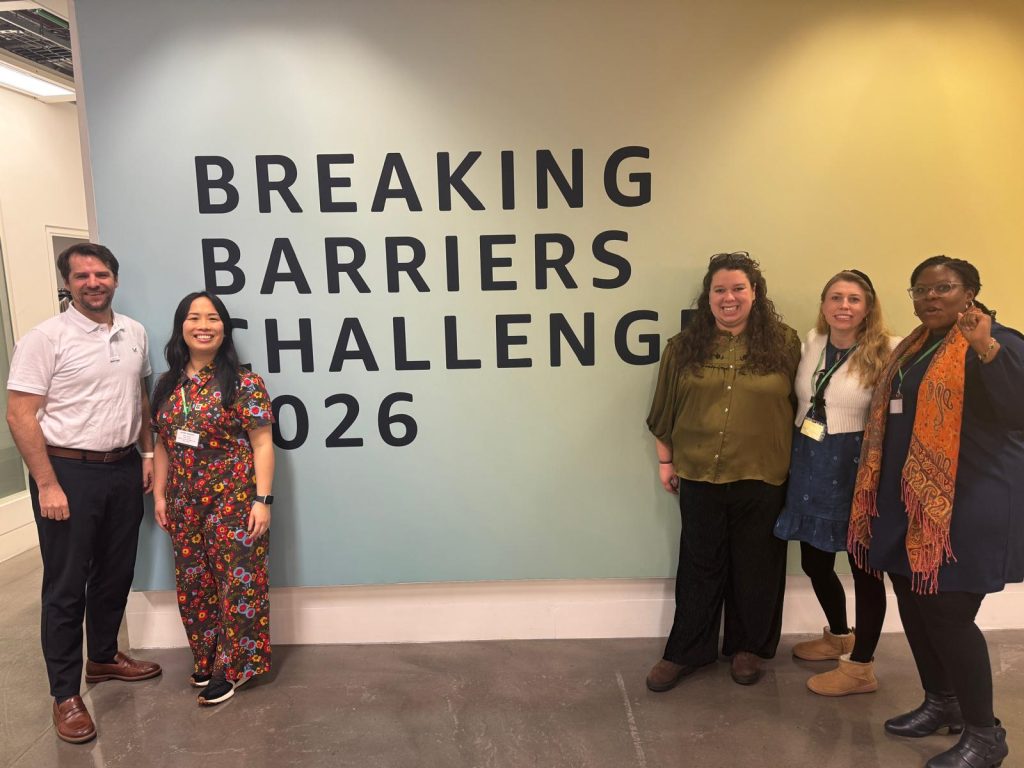 Five people standing and smiling in front of a wall with large text that reads “Breaking Barriers Challenge 2026.” The group represents "From barriers to breakthroughs: HLA at the AWS hackathon 2026" in a modern indoor setting.