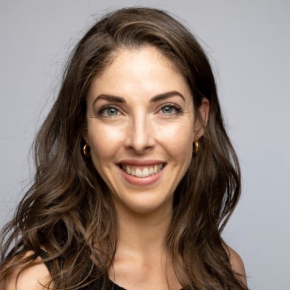 A woman with long brown hair and blue eyes smiles at the camera. She is wearing gold hoop earrings and a black top, ready to discuss the Humanitarian AI January 2026 pulse survey webinar against a light gray background.