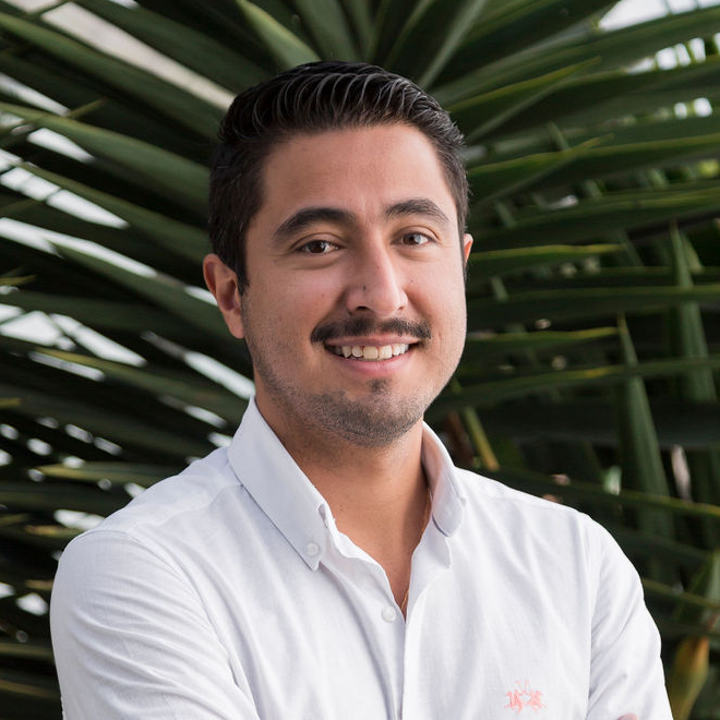 A man with short dark hair and a trimmed mustache and beard, wearing a white button-up shirt, stands smiling with arms crossed in front of large green palm leaves, ready to join the Humanitarian AI January 2026 pulse survey webinar.
