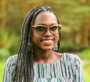 A woman with long braided hair and glasses smiles outdoors, wearing a patterned top in green, black, and white designs. She enjoys fresh air as she prepares to join the Humanitarian AI January 2026 pulse survey webinar. Blurred greenery fills the background.