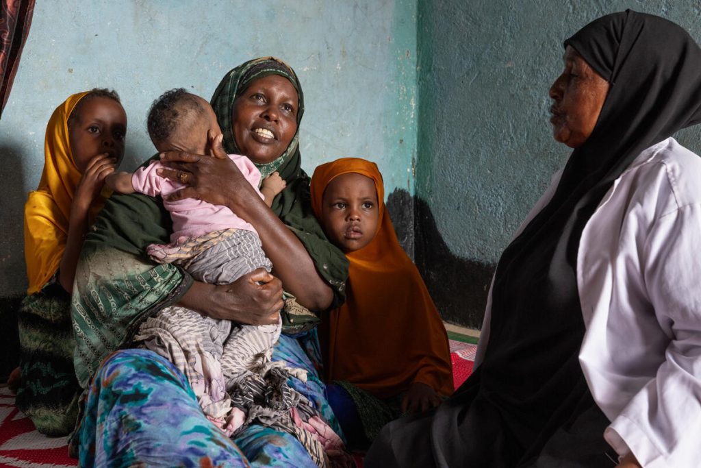 A woman in a green headscarf smiles while holding a baby, sitting beside two young children in yellow headscarves and an older woman in a white coat, all sitting on a red and white mat indoors.