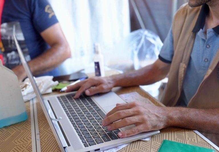 A person is typing on a laptop at a table, with another person sitting nearby. Papers, a green folder, and a bottle of hand sanitizer are also on the table. Only their hands and part of their arms are visible.