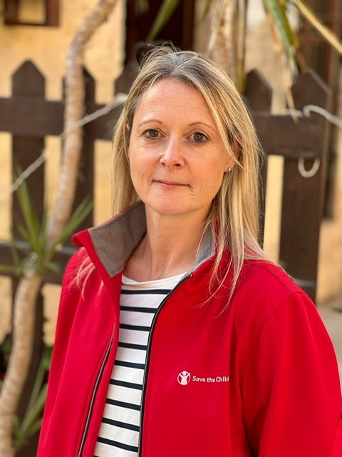 A woman with blonde hair, wearing a red Save the Children jacket over a black-and-white striped shirt, stands outdoors in front of a wooden fence and green plants.