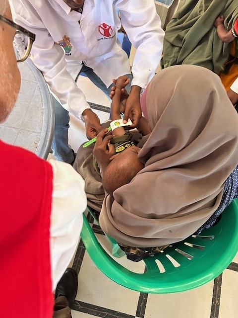 A health worker measures the upper arm circumference of a small child held by a woman in a headscarf, checking for malnutrition. The scene takes place indoors with others nearby.