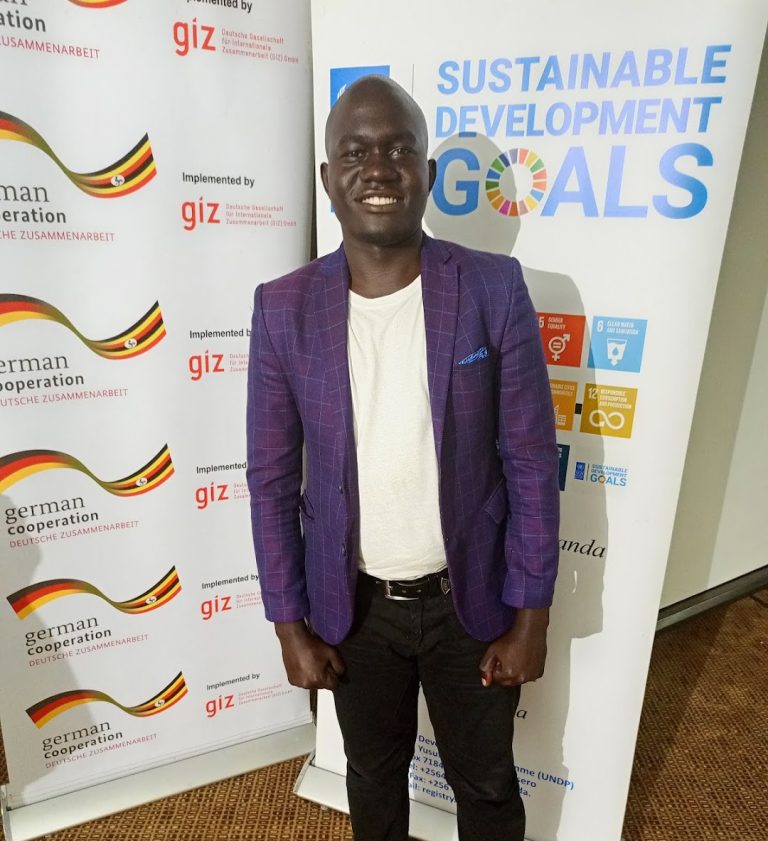 A man in a purple blazer, white shirt, black pants, and bright orange shoes stands smiling in front of banners for the Sustainable Development Goals and German Cooperation.