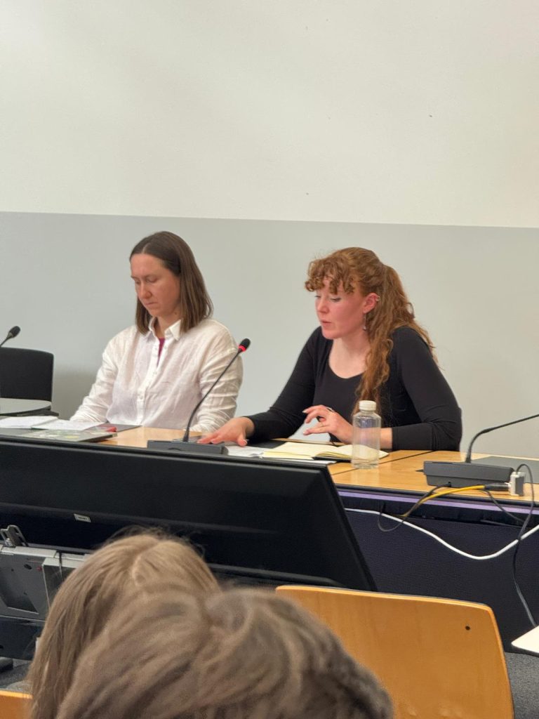 Two people sit at a conference table with microphones, papers, and a water bottle in front of them, appearing to speak or present to an audience in a formal setting.