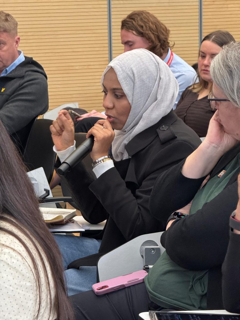 A woman wearing a light gray hijab and black coat speaks into a microphone during a meeting, surrounded by other attentive participants seated in a conference room.